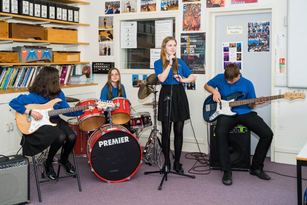 Students playing as part of a band during a music lesson, featuring a guitar, bass, drums and vocals.